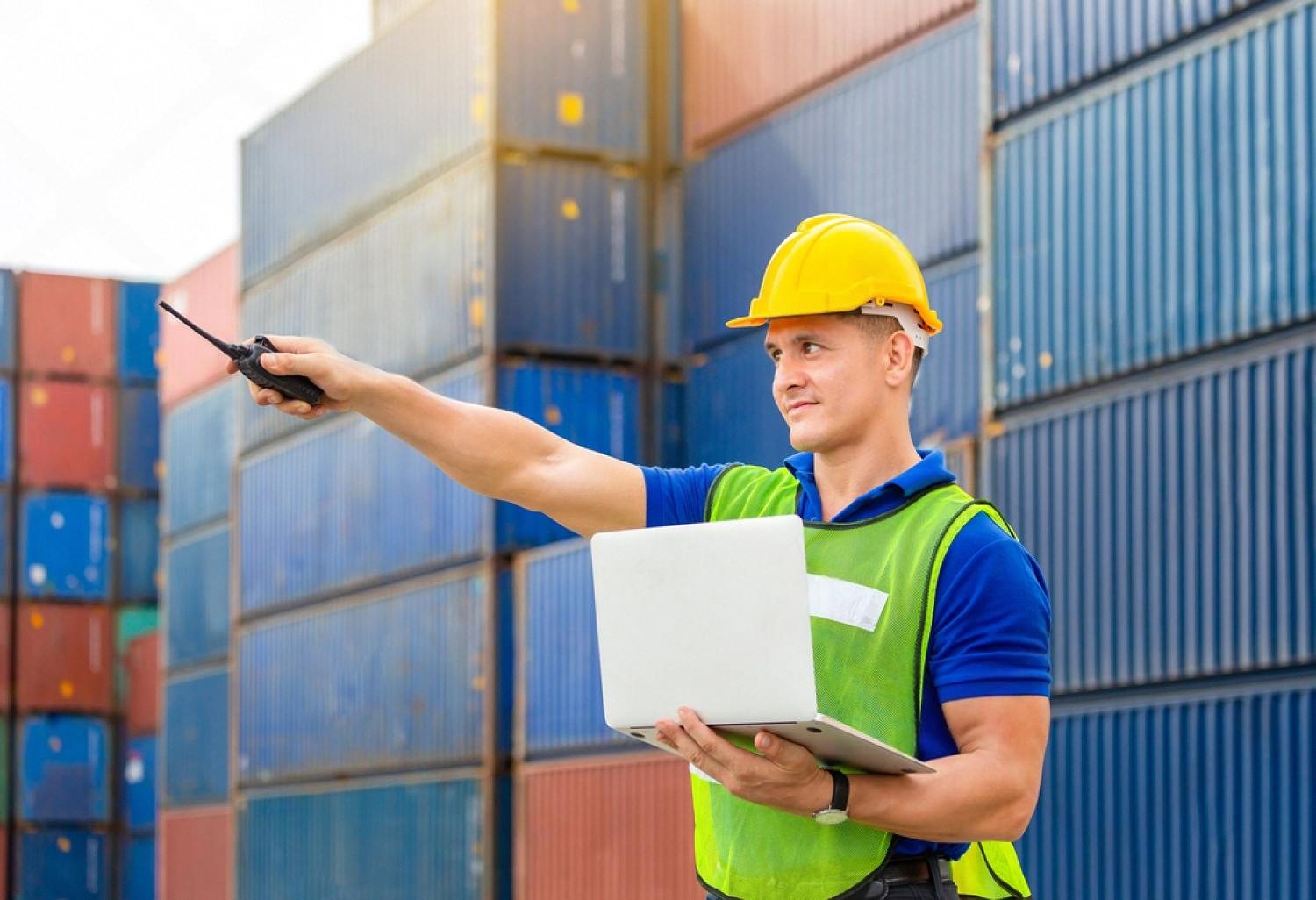 Port operator in a hard hat using a laptop and radio in front of stacked shipping containers at a UAE terminal